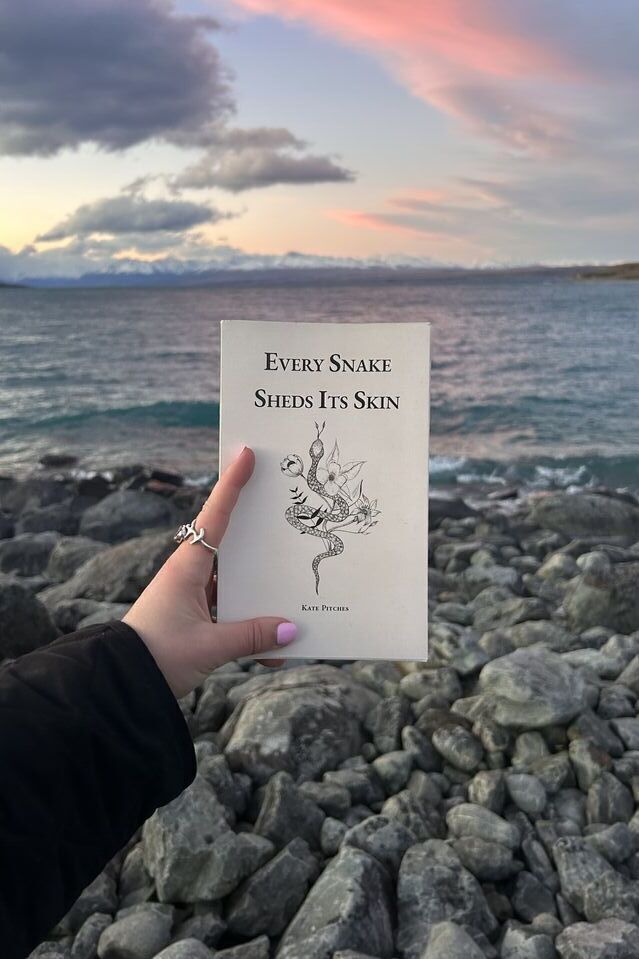A hand holding a cream paperback, Every Snake Sheds Its Skin by Kate Pitches, its cover a line drawing of a snake among flowers, in front of a lake and snow-capped mountains at dusk.